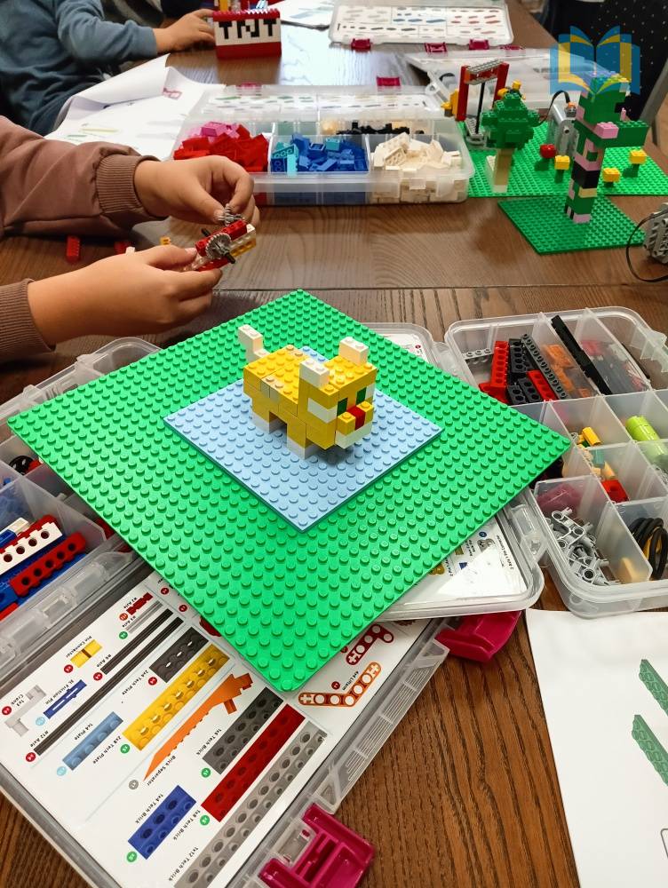 Zdjęcie: Na stole znajdują się pudełka z klockami Lego. Dzieci układają z nich różne modele. Na środku zdjęcia żółty kotek z klocków na zielonej podstawce.